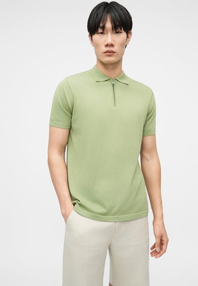 Jeune homme portant un polo à manches courtes vert clair avec une fermeture éclair à l'avant et un pantalon beige clair, debout devant un fond blanc uni.