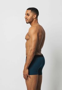 Türkise Boxershorts mit schwarzem Bund, die ein enges Design und eine glatte Textur aufweisen, seitlich auf einem nackten Oberkörper gezeigt.