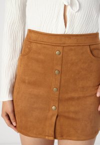 Mini-jupe en daim marron avec cinq boutons en métal sur le devant, comportant deux poches latérales et une fente à l'avant pour plus de détail.