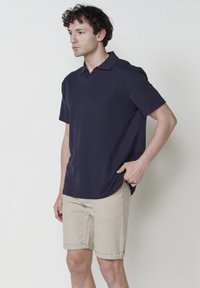 Polo à manches courtes bleu marine en tissu texturé avec un col classique, associé à un short beige retroussé. Logo discret sur la poitrine.
