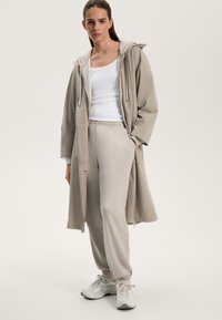 Beige pelerína s kapucí a spadlými rameny přes bílý tílko a světle šedé tepláky. Tenisky s akcenty dotvářejí outfit.