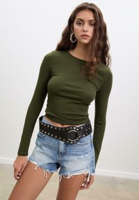 Olivengrünes Langarmoberteil, kombiniert mit ausgewaschenen Denim-Shorts. Schwarzer Gürtel mit runder Schnalle und silbernen Nieten. Neutraler Hintergrund.