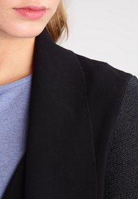 Czarna kurtka z teksturowanym, dzianinowym rękawem, kontrastująca z gładkim materiałem. Pod spodem widoczna jest niebieska bluzka z okrągłym dekoltem.