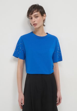 Donna con capelli corti che indossa una maglietta blu con borchie argentate sulle maniche e pantaloni neri a vita alta, in piedi contro un muro bianco.