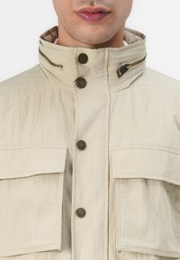 Veste légère beige avec un col montant, deux grandes poches poitrine et des détails en bronze contrastants. Elle dispose d'une fermeture éclair latérale et d'une fermeture à bouton.