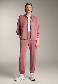 Ružový športový trenčkot so zipsom a kapucňou, ktorý má vrecká, spolu s ladiacimi úzkymi nohavicami s elastickým pásom a bielymi teniskami.