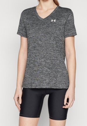 Vrouw draagt een grijs, kortærmlig sportshirt met V-hals en wit logo, en zwarte fietsshort, staand tegen een effen achtergrond.