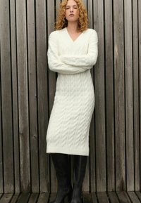 Rochie tricotată cu mâneci lungi, de culoare albă, cu decolteu în V și tiv ribbit, purtată cu cizme negre până la genunchi, pe un fundal din lemn.