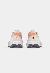 Baskets de sport blanches avec une tige texturée gris clair, des accents orange et une semelle moelleuse avec un crampon noir. Logo affiché sur le talon.