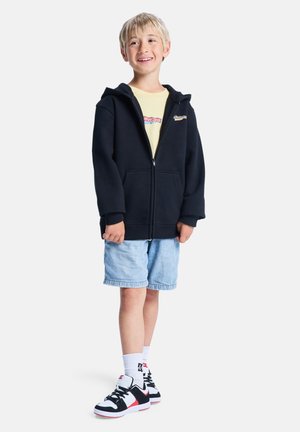 Czarna bluza z kapturem zapinana na zamek, noszona na jasnosłonecznej żółtej koszulce. Para jasnych szortów jeansowych i czarne trampki z czerwonymi akcentami.