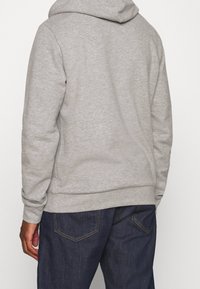 Sudadera con capucha gris de tela texturizada, puños y dobladillo acanalados, vista desde atrás, combinada con vaqueros oscuros que tienen bolsillos visibles.