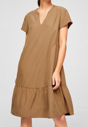Vrouw draagt een loszittende, lichtbruine jurk met korte mouwen, een V-hals en een gelaagde zoom, staand tegen een effen achtergrond.