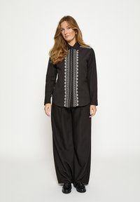 Čierna košeľa s dlhým rukávom a golierom na gombíky, s bielymi vyšívanými detailmi po prednej strane, v kombinácii s voľnými čiernymi nohavicami.