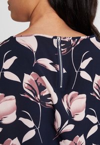 Blusa blu navy con grandi motivi floreali rosa, scollatura rotonda e zip argentata visibile sul retro. Tessuto liscio e leggero.