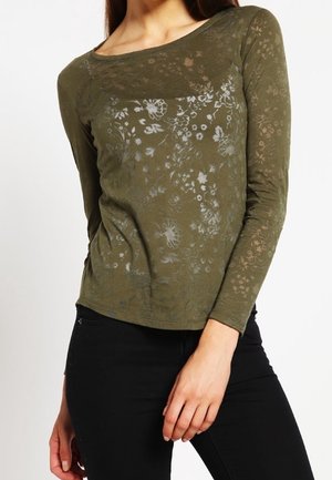 Maglietta a maniche lunghe verde oliva con un motivo floreale testurizzato. Presenta un collo a barchetta e un design aderente. Indossata con pantaloni neri.