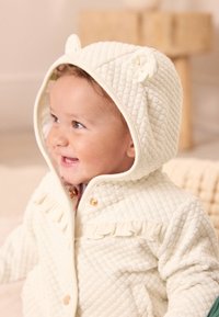 Manteau pour bébé matelassé crème avec une capuche à oreilles d'ourson, bordure à volants et boutons pression dorés. Texture douce, design chaud et palette de couleurs neutres.