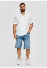 Witte poloshirt met een groen palmbomenpatroon, gecombineerd met blauwe denim shorts. Model draagt witte sneakers en staat met een ontspannen houding.