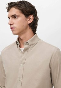 Camisa de botones beige con cuello extensible, textura suave y un bolsillo en el pecho. Presenta botones perlados y un diseño de corte relajado.
