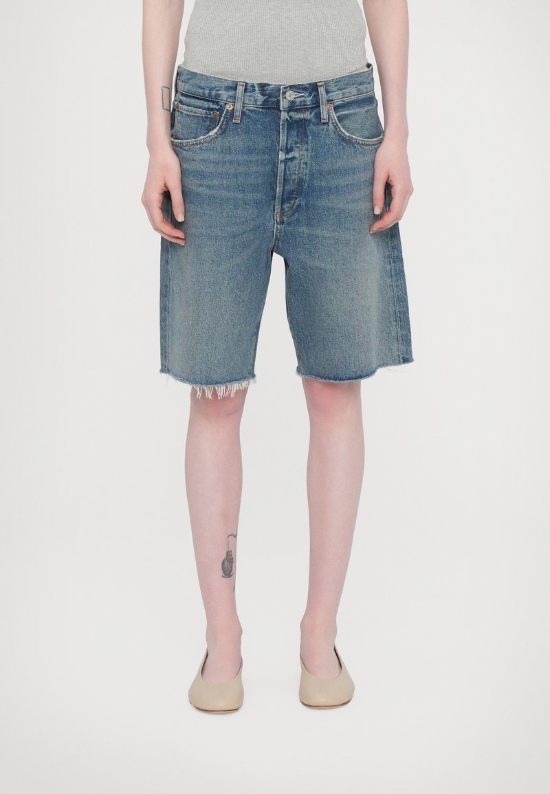 Personne portant un débardeur gris, un short en jean déchiré à l'ourlet arrivant au genou, des chaussures plates beiges, avec un tatouage sur la jambe gauche inférieure.
