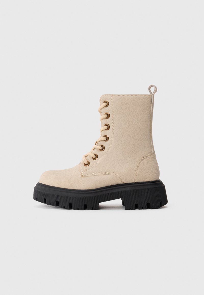 Beige lær kampstøvle med tyk sort sål og beige snørebånd, set fra ydersiden på en ensfarvet lys baggrund.