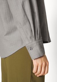 Camicia grigia a spina di pesce con maniche lunghe e polsini abbottonati, abbinata a pantaloni verde oliva. Il tessuto appare morbido e strutturato.
