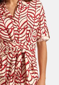 Rode en crèmekleurige jurk met een bladpatroon, knoopsluiting aan de voorkant, korte omgeslagen mouwen en een strikdetail in de taille. Soepele stoftextuur.