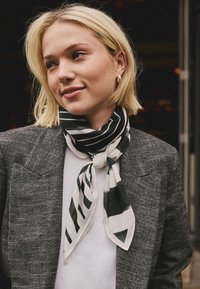 Schwarz-weiß gestreifter Seidenschal, am Hals gebunden, mit leichter Textur und Knoten-Detail, getragen zu einem grauen Blazer.