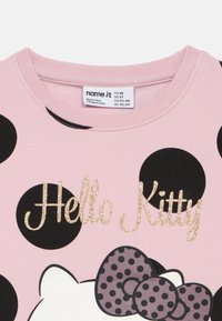 Sweatshirt rosa com bolinhas pretas, apresentando "Hello Kitty" em texto dourado brilhante e um padrão de laços em cinza com bolinhas pretas.