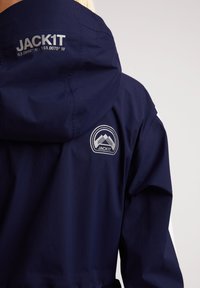 Chaqueta impermeable color azul marino con capucha, logo "JACK1T" y gráfico de montaña en la parte posterior. Textura suave con detalles en acento blanco.