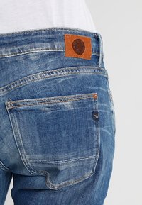 Jeans de mezclilla azul con un parche de cuero marrón en la parte trasera de la cintura, que cuentan con bolsillos, detalles de desgaste y costuras en contraste.