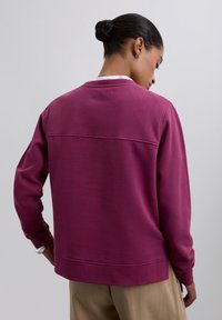 Mujer con el cabello recogido en un moño, vistiendo una blusa de manga larga morada y pantalones beiges, girada ligeramente de lado frente a un fondo neutral.