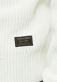 Maglione bianco a maglia con texture a coste. Presenta un'etichetta nera e oro con "JACK & JONES" e ulteriori dettagli del brand.