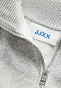 Chaqueta gris con cremallera, cuenta con un cuello de canalé, un forro interior suave, cremallera metálica y una etiqueta blanca con texto azul "JJXX".