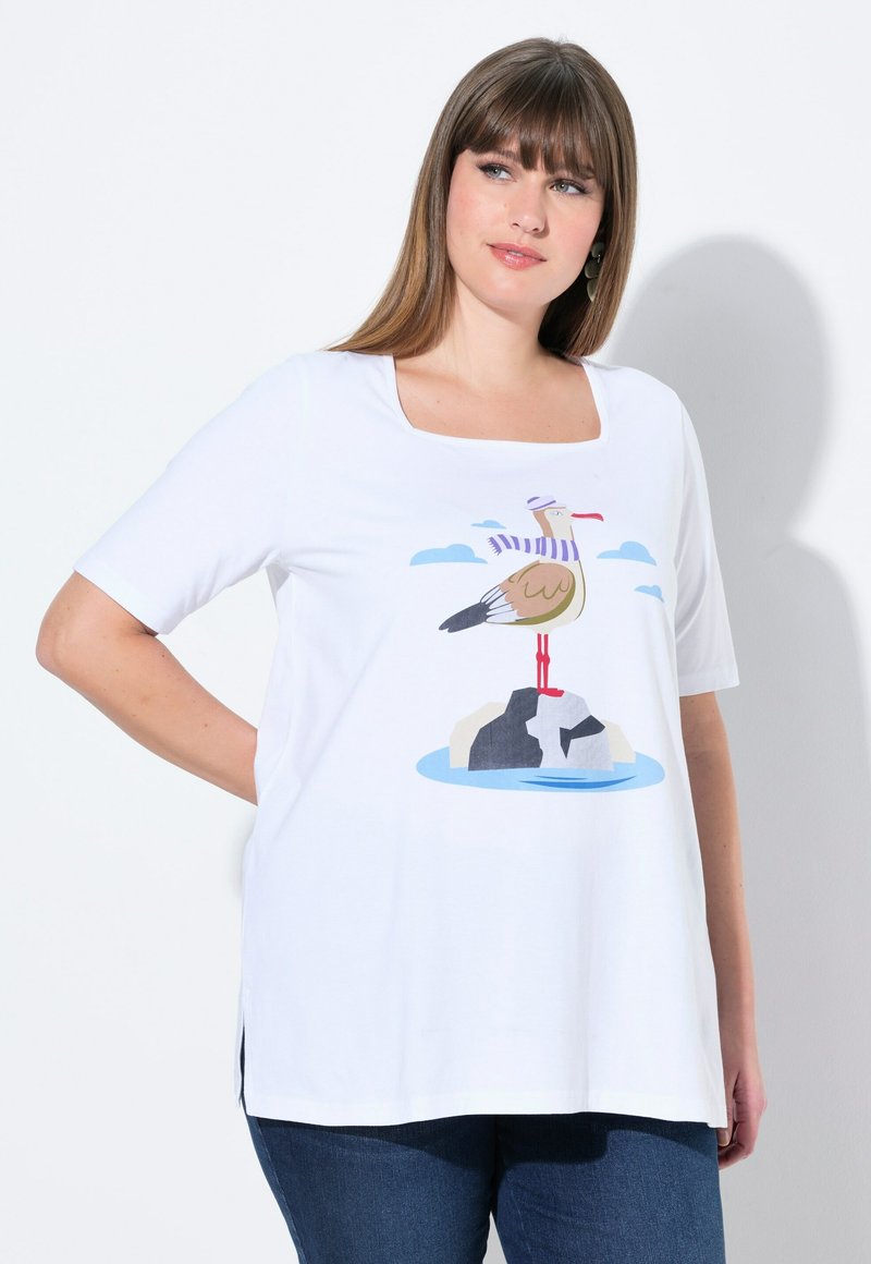 T-shirt en coton blanc avec un décolleté carré. Présente un graphique coloré d'un oiseau sur des rochers avec des nuages au-dessus, des manches courtes et des fentes sur les côtés.