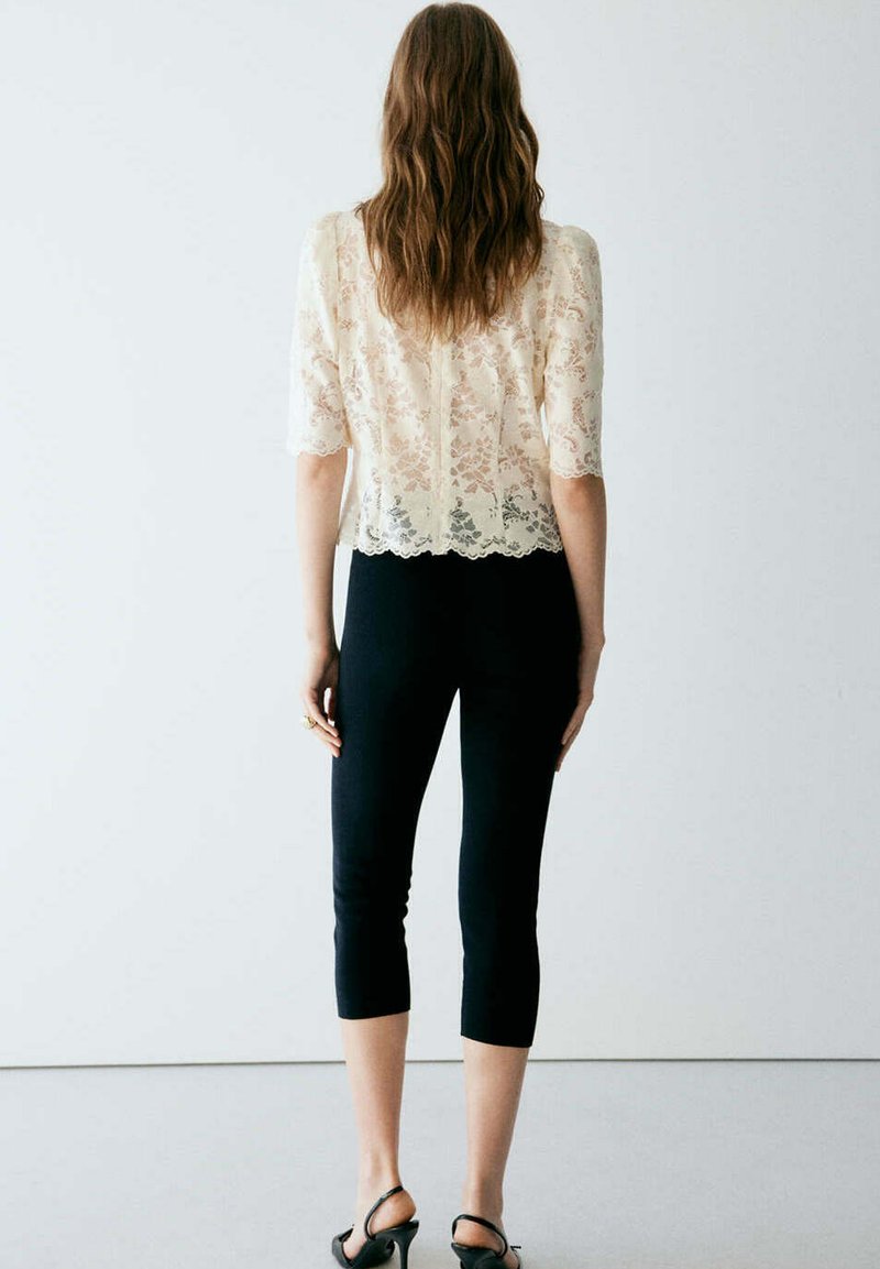 Femme aux longs cheveux bruns porte un haut en dentelle crème à manches coudes, un pantalon court noir et des talons noirs, debout de dos sur un fond uni.