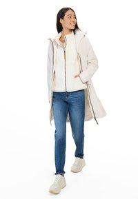 Hellbeige wasserfester Mantel mit Kapuze, über einer cremefarbenen Fleecejacke getragen, kombiniert mit blauen Jeans und beigen Sneakers.