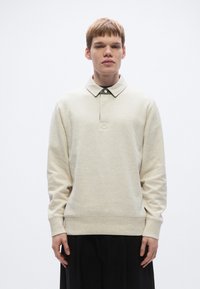 Lysebeige polosweatshirt med krave og knaplukning, som har lange ærmer og en struktureret overflade. Model iført sorte bukser.