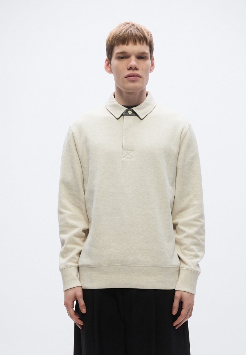 Lysebeige polosweatshirt med krave og knaplukning, som har lange ærmer og en struktureret overflade. Model iført sorte bukser.