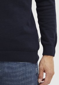 Marineblauer gestrickter Pullover mit gerippten Bündchen und Saum, der ein kleines strukturiertes Etikett an der Seite aufweist. Das Material wirkt weich und formschlüssig.