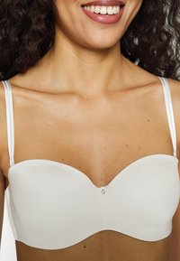 Witte strapless bh van gladde stof, met een gevormde pasvorm en een kleine decoratieve sluiting in het midden. Verstelbare, dunne bandjes.