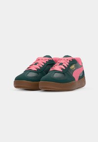 Green suede sneakers with pink laces and accents, featuring a smooth texture and a rubber sole with a ridged pattern. Gold branding on side.