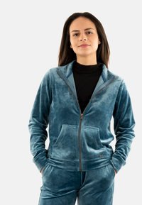 Giacca zip-up in velluto color turchese, con colletto alto, maniche lunghe e tasche frontali. Tessuto liscio con una leggera lucentezza.