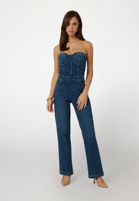 Džínsový overal s bezramenným telom, priliehavým pásom, širokými nohavicami, tmavomodrou farbou a prednými vreckami. Obsahuje švy pozdĺž tela.