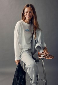 Cămașă albă cu mâneci lungi cu logo-ul "CHEVROLET", pantaloni de trening gri și încălțări portocalii cu imprimeu de leopard, așezat pe un scaun argintiu cu un sacou negru.