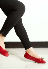 Ballerines en velours rouge avec un bout arrondi et un petit détail de nœud, portées avec des leggings noirs, mettant en avant un design épuré et simple.