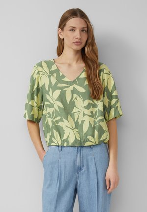 Woman with long hair wearing a green floral V-neck blouse and light blue high-waisted jeans, standing with one hand in pocket.