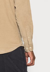 Camisa de pana beige con mangas largas, que presenta puños abotonados y una superficie texturizada. Mano posicionada cerca del dobladillo, revelando un tejido oscuro debajo.