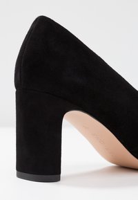 Scarpa nera in suede con tacco alto, a punta arrotondata e sovratessuto texture. Presenta un tacco a blocco robusto e una fodera interna liscia.