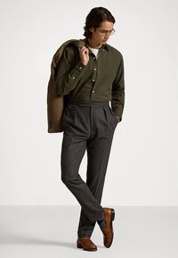 Oliwkowa, zapinana na guziki koszula, szare plisowane spodnie i brązowe skórzane buty. Teksturowana brązowa kurtka zarzucona na ramię.