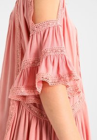 Růžový lehký top z tkaniny s designem studeného ramene, který má vrstvené volánkové rukávy a jemnou krajkovou aplikaci podél okrajů.